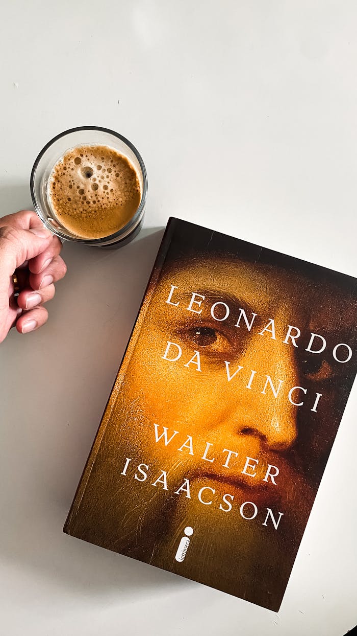 A person enjoys coffee while holding a Leonardo da Vinci book, photographed from above.